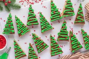 A close-up overhead view of a triangular Rice Krispie treat decorated like a Christmas tree with green frosting and red sprinkles, sitting on a white plate next to a vintage fork and a sprig of pine.