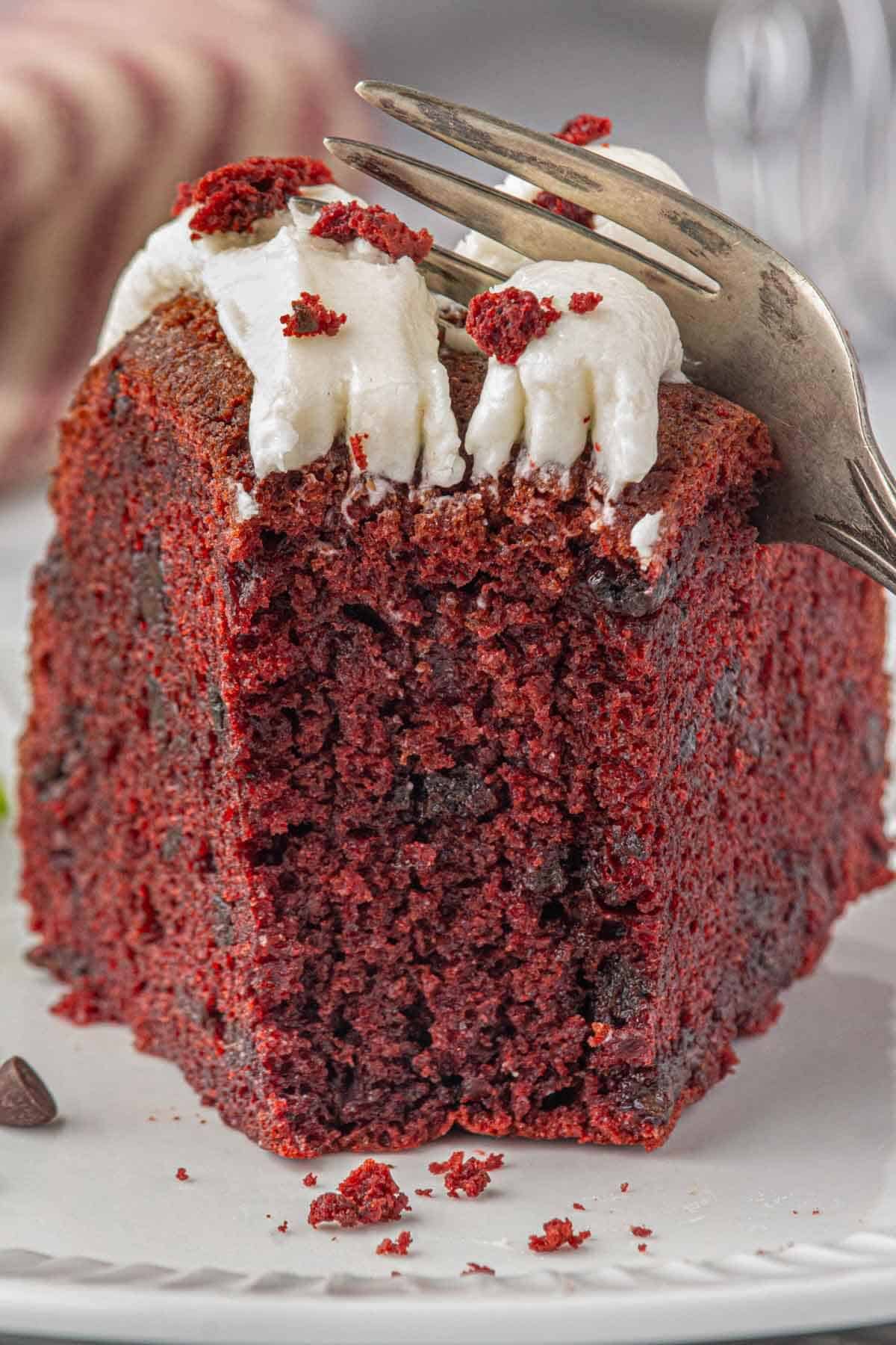 Close-up of a fork cutting into a slice of red velvet Bundt cake with cream cheese frosting and red velvet crumb topping.