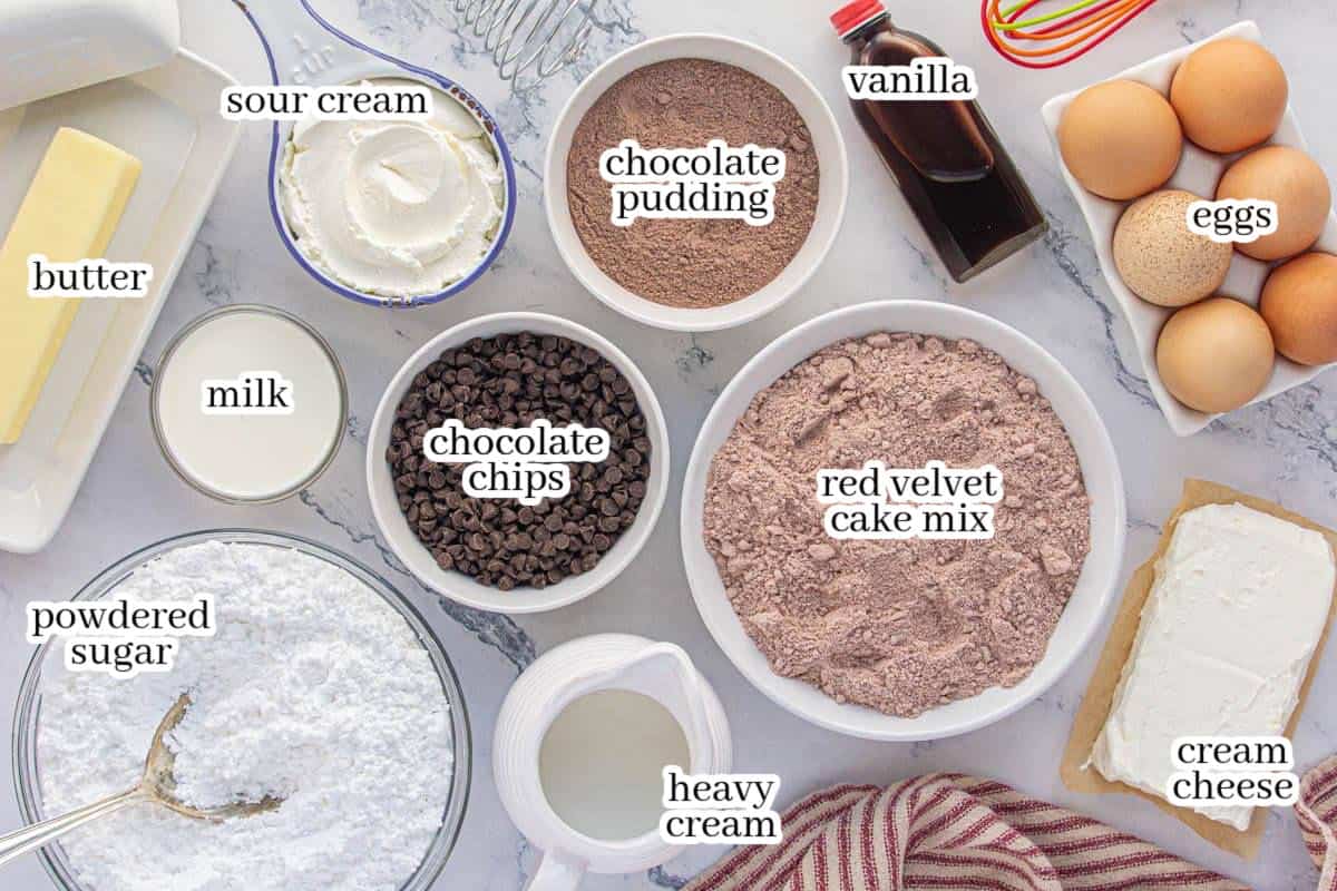 Overhead view of labeled ingredients for Copycat Nothing Bundt Red Velvet Cake, including butter, sour cream, chocolate pudding mix, vanilla extract, eggs, milk, chocolate chips, red velvet cake mix, powdered sugar, heavy cream, and cream cheese.