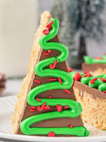 A single Christmas tree Rice Krispie treat standing upright on a plate, with chocolate topping, green frosting piped in a zigzag pattern, and red holiday sprinkles.