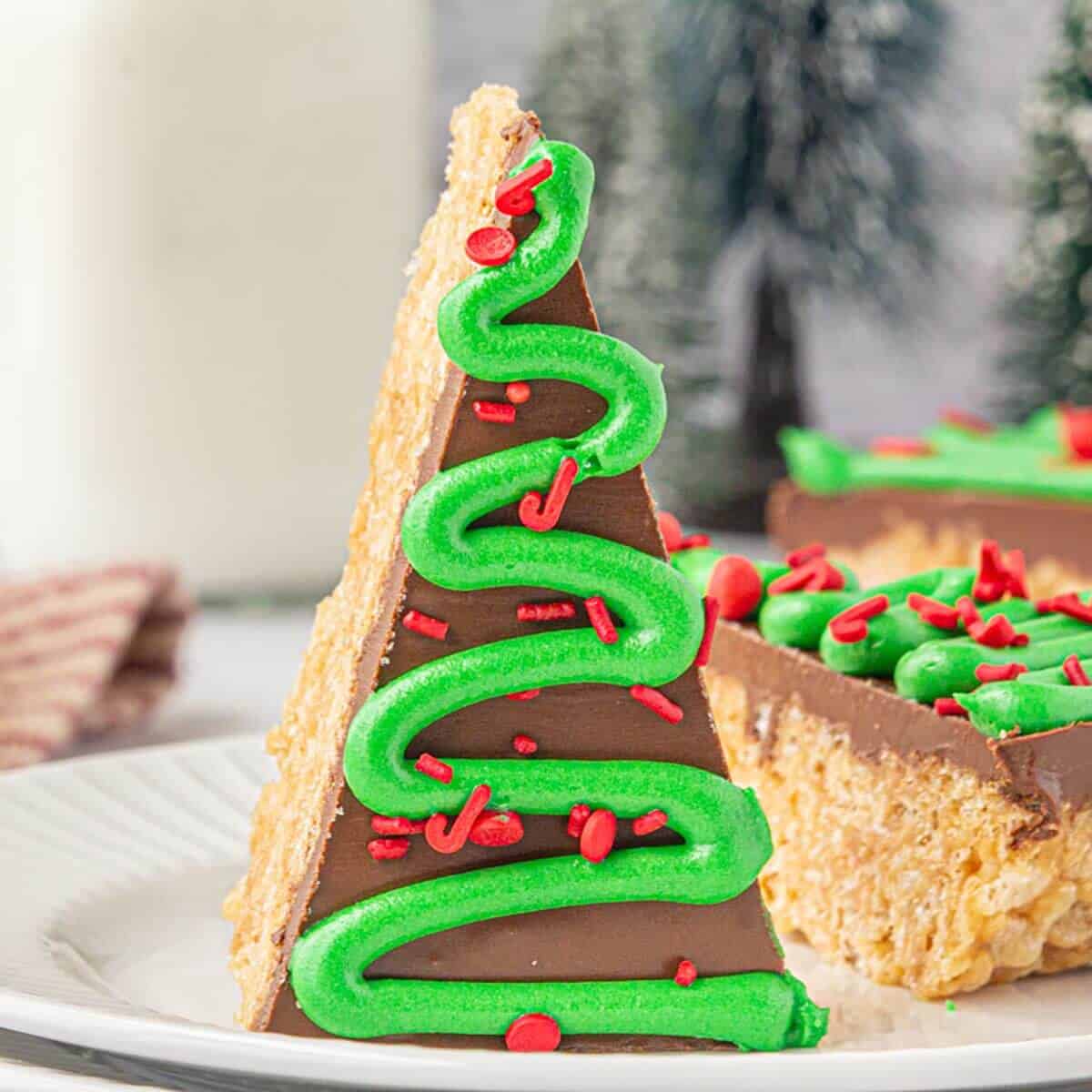 A single Christmas tree Rice Krispie treat standing upright on a plate, with chocolate topping, green frosting piped in a zigzag pattern, and red holiday sprinkles.