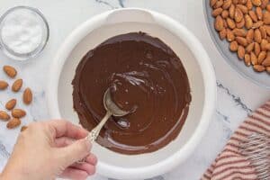 Melted chocolate stirred with a silver spoon in a white mixing bowl, with toasted almonds and sea salt nearby.