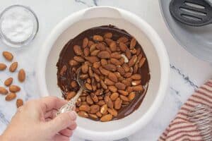 Toasted almonds being stirred into a bowl of melted chocolate with a silver spoon, surrounded by scattered almonds and sea salt.