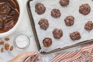 Chocolate almond mixture being scooped onto a parchment-lined baking sheet using a cookie scoop, with finished clusters sprinkled with sea salt.