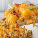 Gooey serving of Tater Tot Bacon Cheeseburger Casserole being lifted from a white baking dish, with melted cheese stretching and crispy bacon on top.