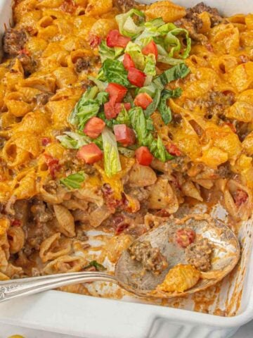 Baked cheeseburger pasta casserole in a white dish, topped with shredded lettuce and tomatoes, with a large serving spoon in the corner.