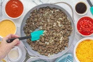 Ground beef and diced onions being sautéed in a large skillet, surrounded by bowls of tomato soup, cheddar cheese soup, Rotel, Worcestershire sauce, evaporated milk, and shredded cheese.