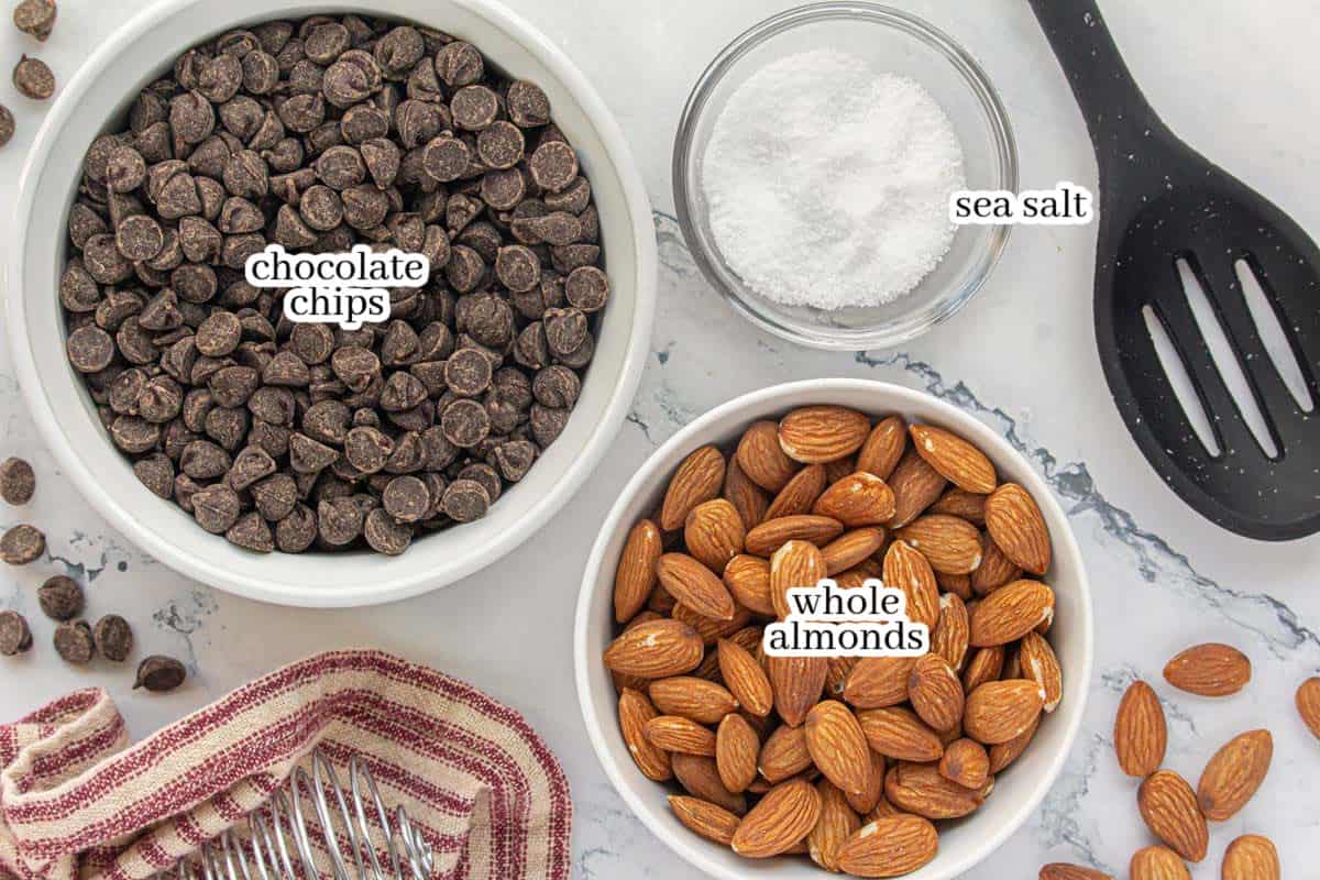 Overhead view of ingredients for candy recipes: a bowl of chocolate chips, a bowl of whole almonds, and a small bowl of sea salt on a marble surface with a striped towel and whisk.