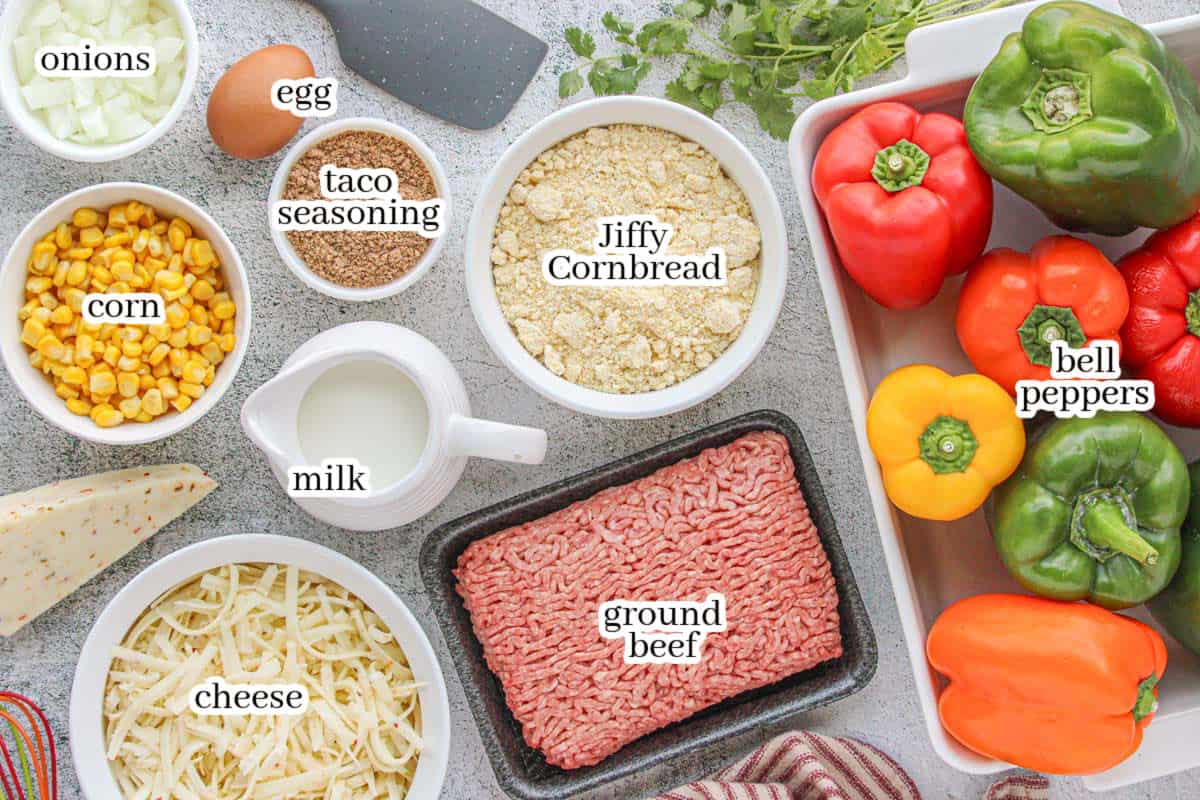 Overhead view of Tex-Mex stuffed pepper ingredients, including bell peppers, Jiffy cornbread mix, ground beef, shredded cheese, taco seasoning, corn, onions, milk, and an egg, arranged around a white baking dish.