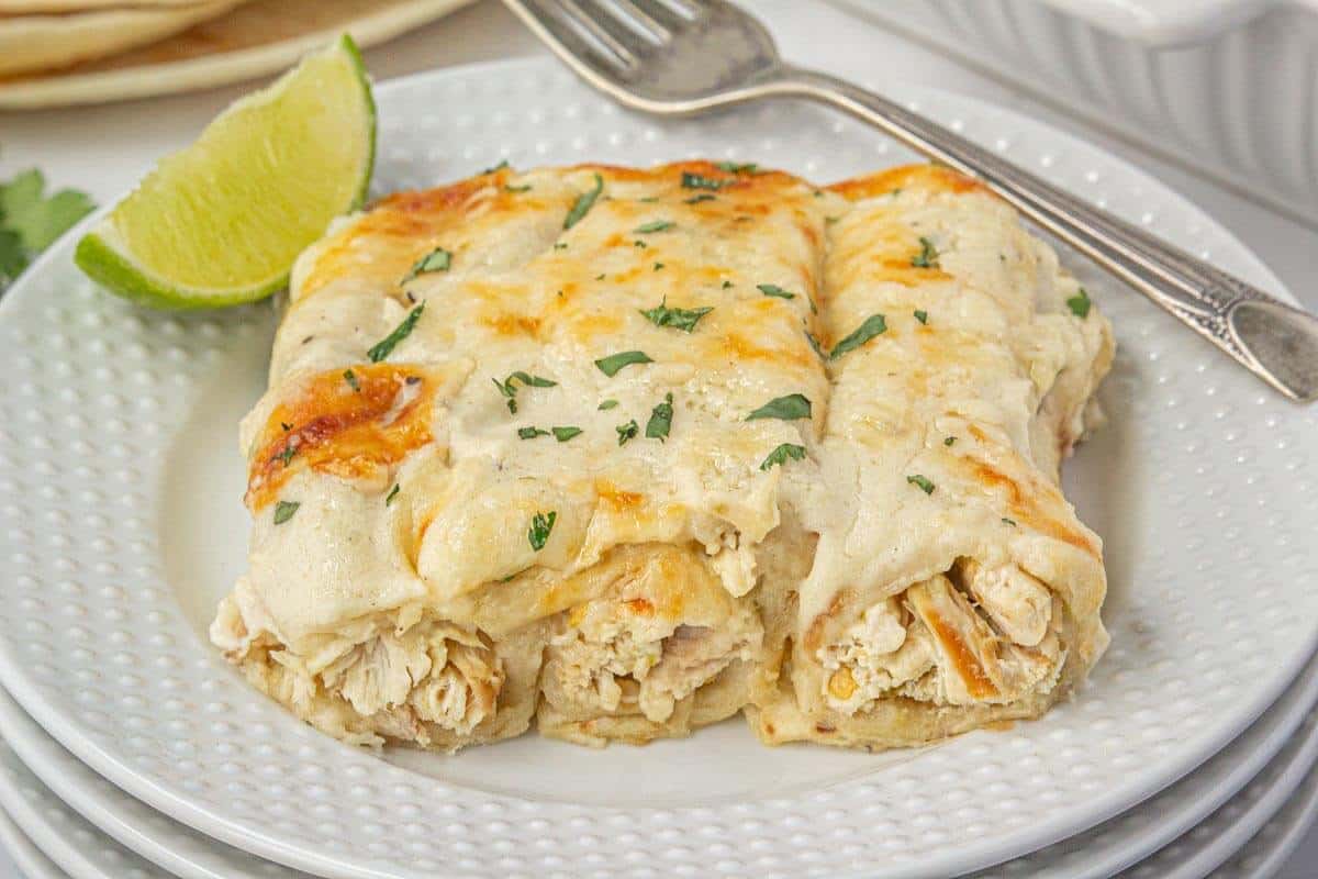 Three creamy chicken enchiladas on a white plate, smothered in melted cheese and sour cream sauce, served with a lime wedge for a pop of color and flavor.