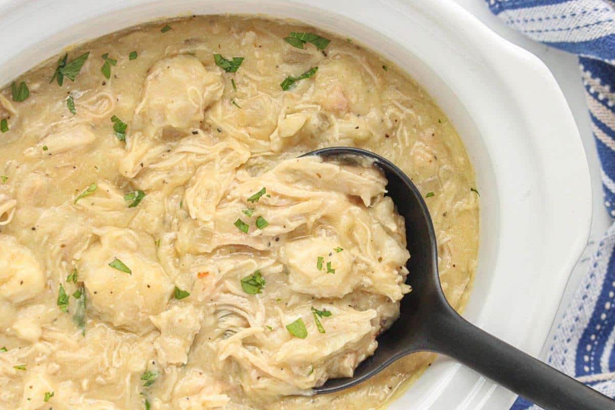 Slow cooker filled with creamy chicken and dumplings, garnished with fresh parsley, with a black serving ladle ready to scoop.