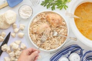 Cooked chicken being shredded with forks on a plate, with a slow cooker of broth and chopped biscuit dough pieces on the side.