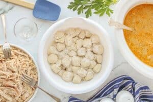 Biscuit dough pieces coated in flour in a white bowl next to shredded chicken and a crockpot of broth.
