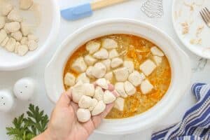 Hand holding floured biscuit dough pieces over a crockpot filled with simmering chicken broth, ready to be dropped in as dumplings.