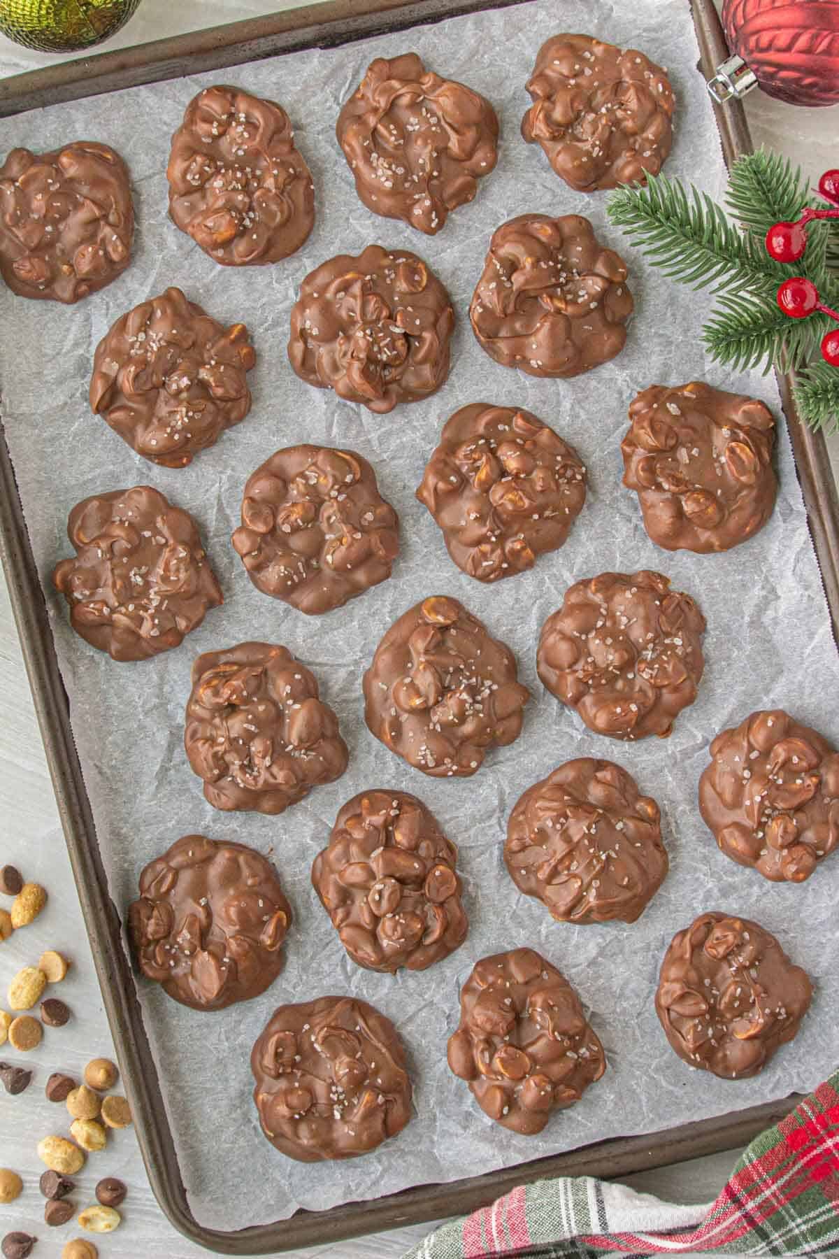 Baking sheet lined with parchment paper holding rows of homemade chocolate peanut clusters, decorated with holiday greenery and ornaments.