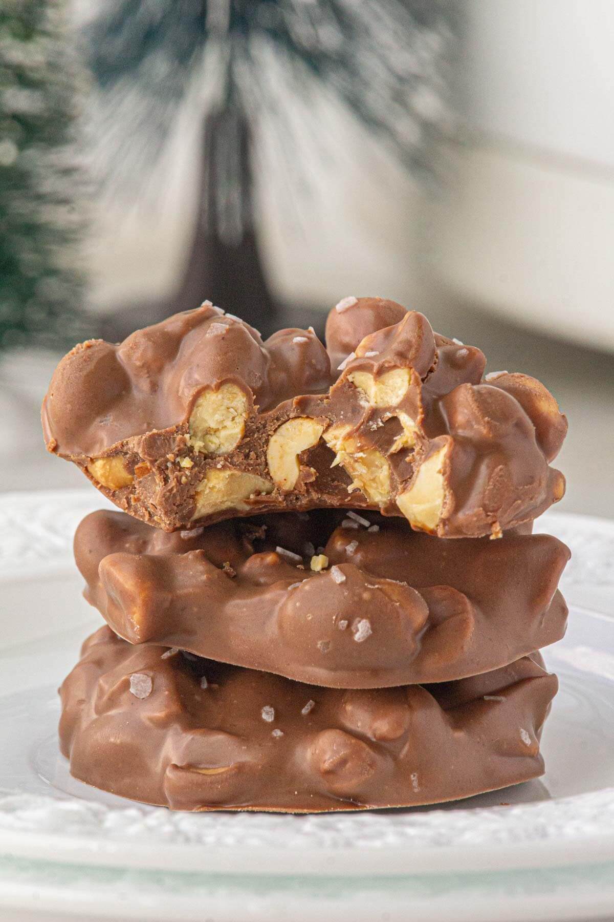 Stack of three chocolate peanut clusters on a white plate, with the top piece broken in half to show the crunchy peanuts inside.