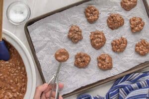 Cookie scoop dropping spoonfuls of melted chocolate peanut clusters onto a parchment-lined baking sheet.