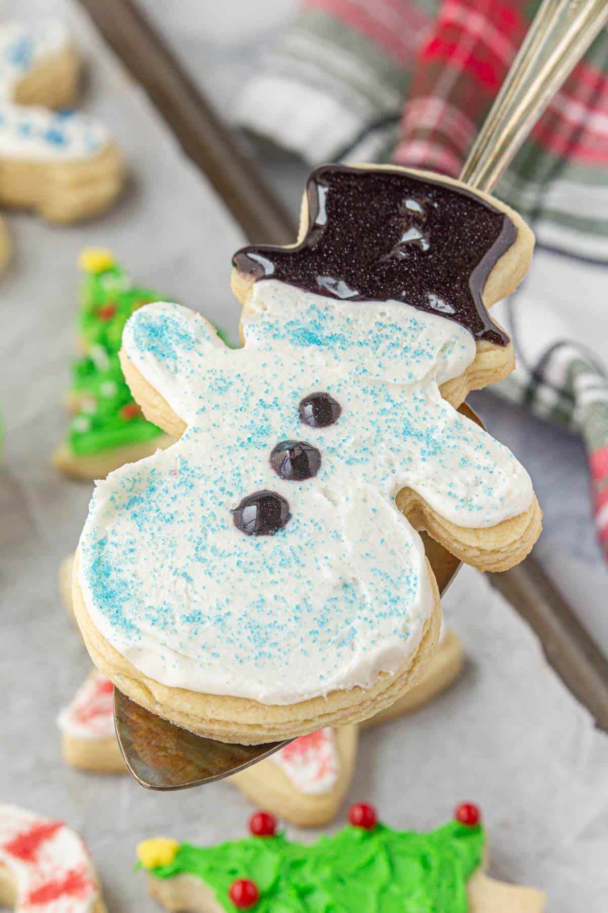 Decorated snowman sugar cookie on a spatula. In the background are more decorated sugar cookies on a baking sheet.