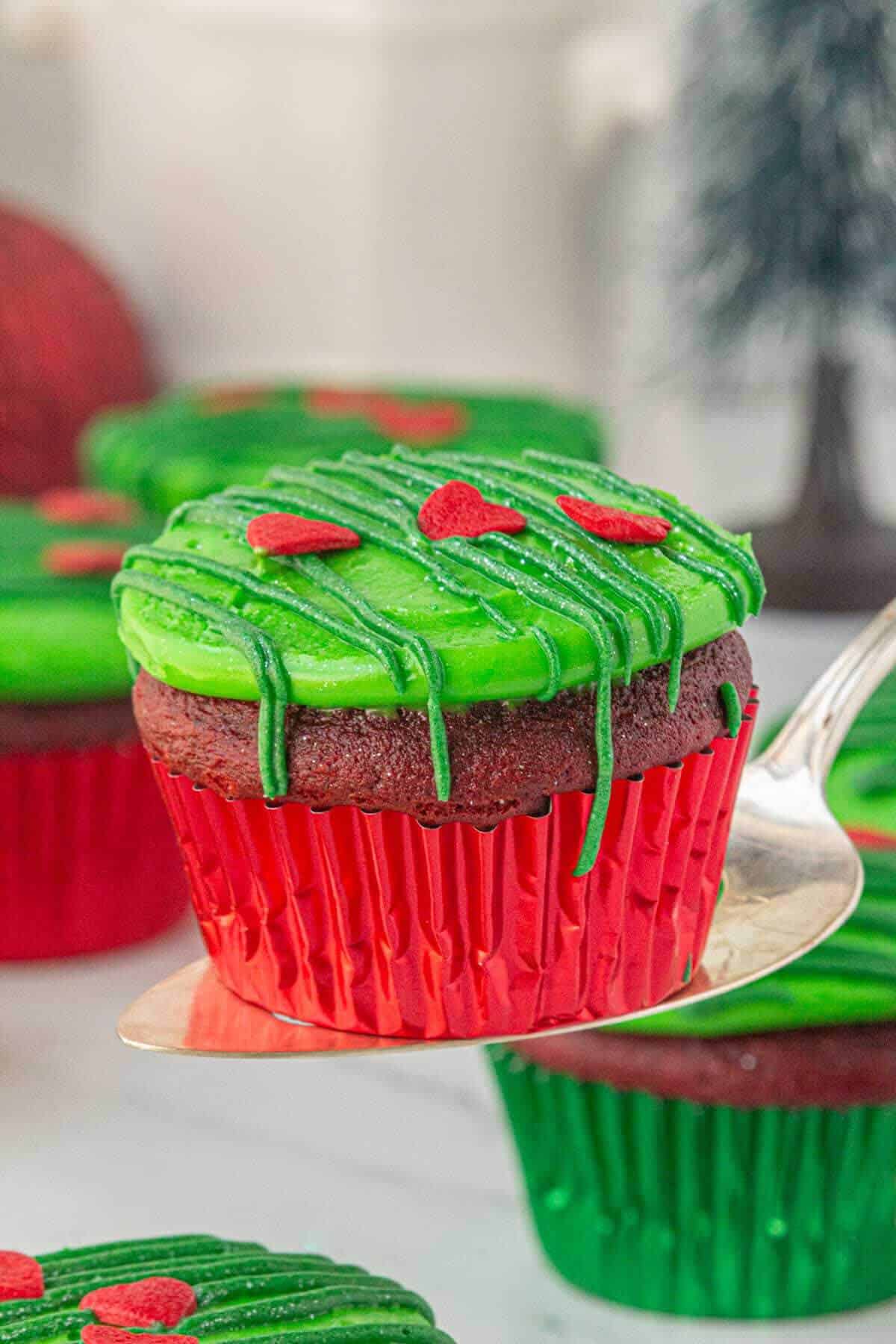 Holiday-themed Grinch Cupcake in a shiny red wrapper, topped with vibrant green frosting, green drizzle, and candy hearts, being held up on a silver spatula.