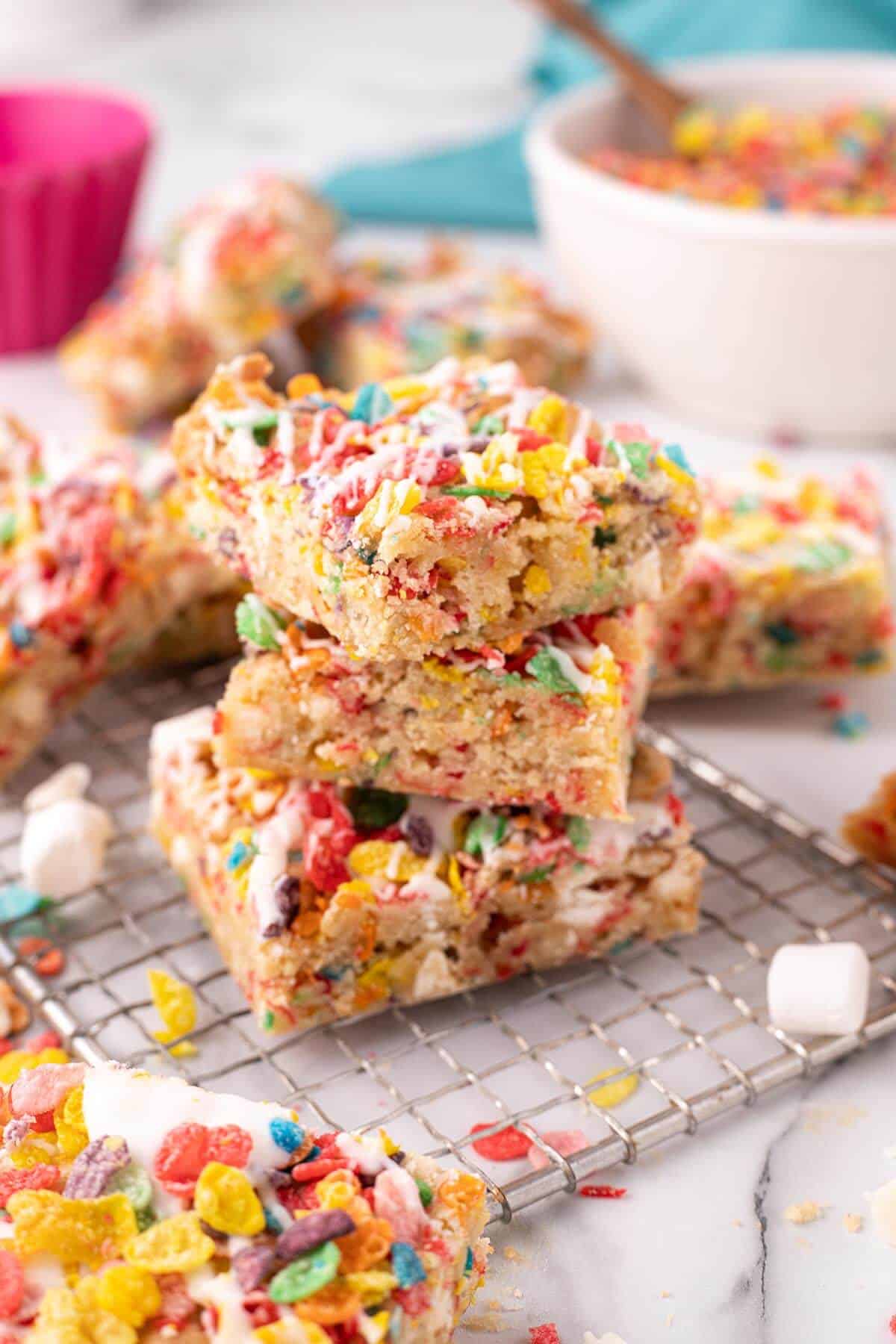Stacked Fruity Pebbles blondies on a cooling rack with a bowl of cereal in the background, showing their chewy, colorful texture.