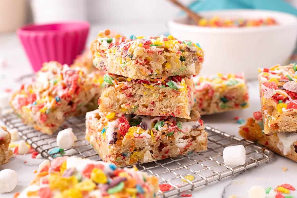 Stack of colorful Fruity Pebbles blondies on a cooling rack, showing chewy centers with bright cereal pieces and a drizzle of white glaze.