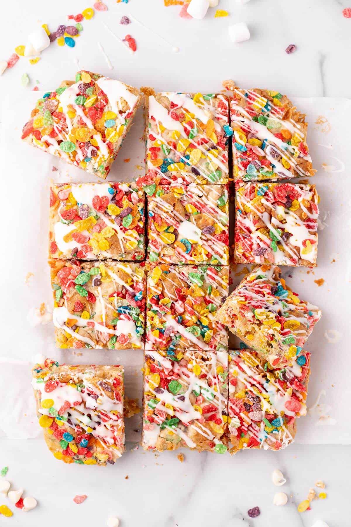 Overhead view of Fruity Pebbles blondies cut into neat squares, topped with vibrant cereal pieces and a drizzle of glaze.