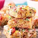 Stack of chewy Fruity Pebbles Blondies on a cooling rack, topped with colorful cereal and white chocolate drizzle, with bold text overlay reading "Fruity Pebbles Blondies" and the Bowl-Me-Over.com URL at the bottom.