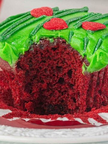 Close-up of a red velvet Grinch Cupcake with a big bite taken out, showing the moist, fluffy center and topped with bright green frosting and red candy hearts.