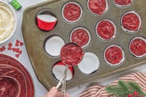 Cupcake liners filled with red cupcake batter in a muffin tin, surrounded by heart sprinkles and food coloring on a holiday-themed workspace.