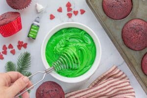 Bright green frosting being whipped in a white bowl with a Danish dough whisk, surrounded by baked cupcakes and red heart sprinkles.