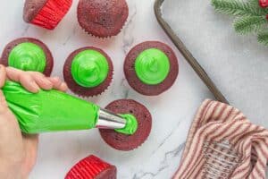 Red velvet cupcakes being piped with vibrant green frosting using a piping bag, with a festive towel and evergreens nearby.