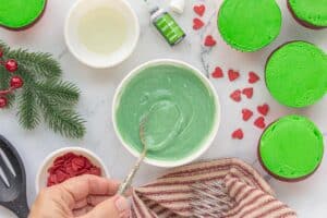 Bowl of green glaze being stirred with a spoon, surrounded by undecorated cupcakes, red heart sprinkles, and green food coloring.
