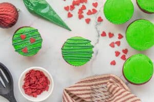 Overhead view of decorated Grinch Cupcakes with green frosting, green drizzle, and red heart sprinkles on a marble surface.