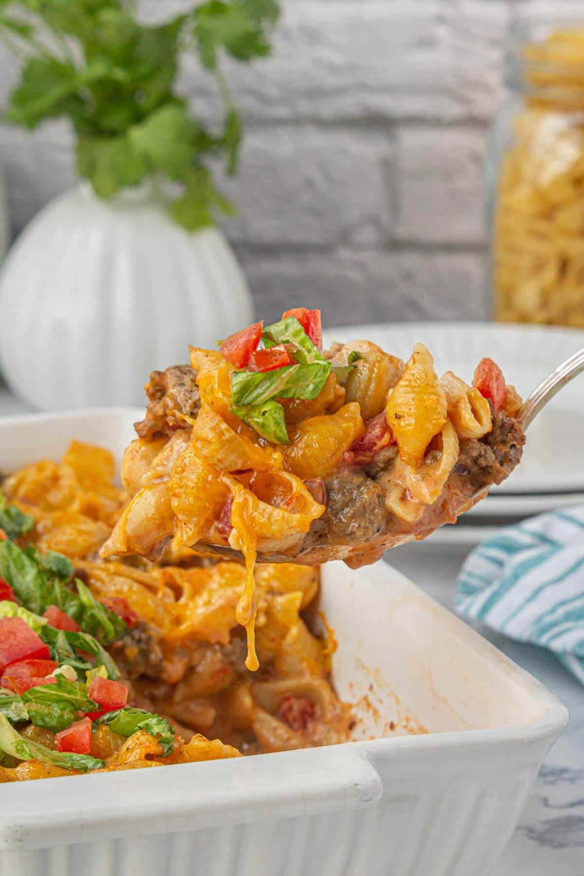 A large spoonful of cheesy cheeseburger pasta casserole lifted from a baking dish, topped with chopped lettuce and diced tomatoes.