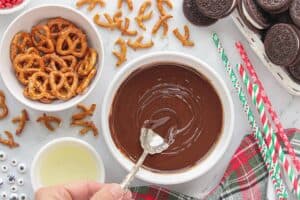 Melted chocolate in a white bowl being stirred by a small spoon. Surrounding the bowl are holiday paper straws, Oreos and pretzels.