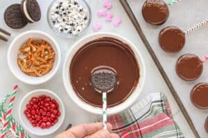 An Oreo cookie being dipped into melted chocolate. Surrounded by candy eyes, mini m&m's and pretzels.