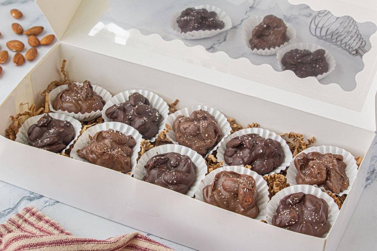 White bakery gift box filled with chocolate almond clusters in paper cups, ready for gifting with shredded paper filler and a clear lid.