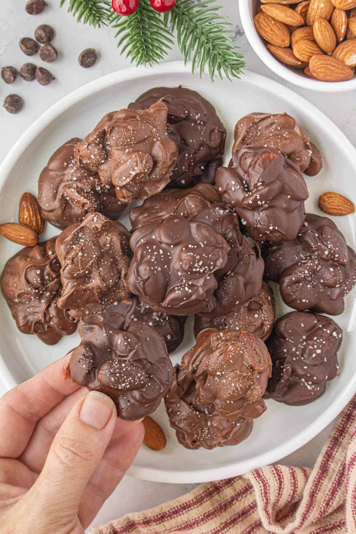 Plate of chocolate almond clusters sprinkled with sea salt, surrounded by holiday greenery, chocolate chips, and whole almonds, with a hand picking one up.