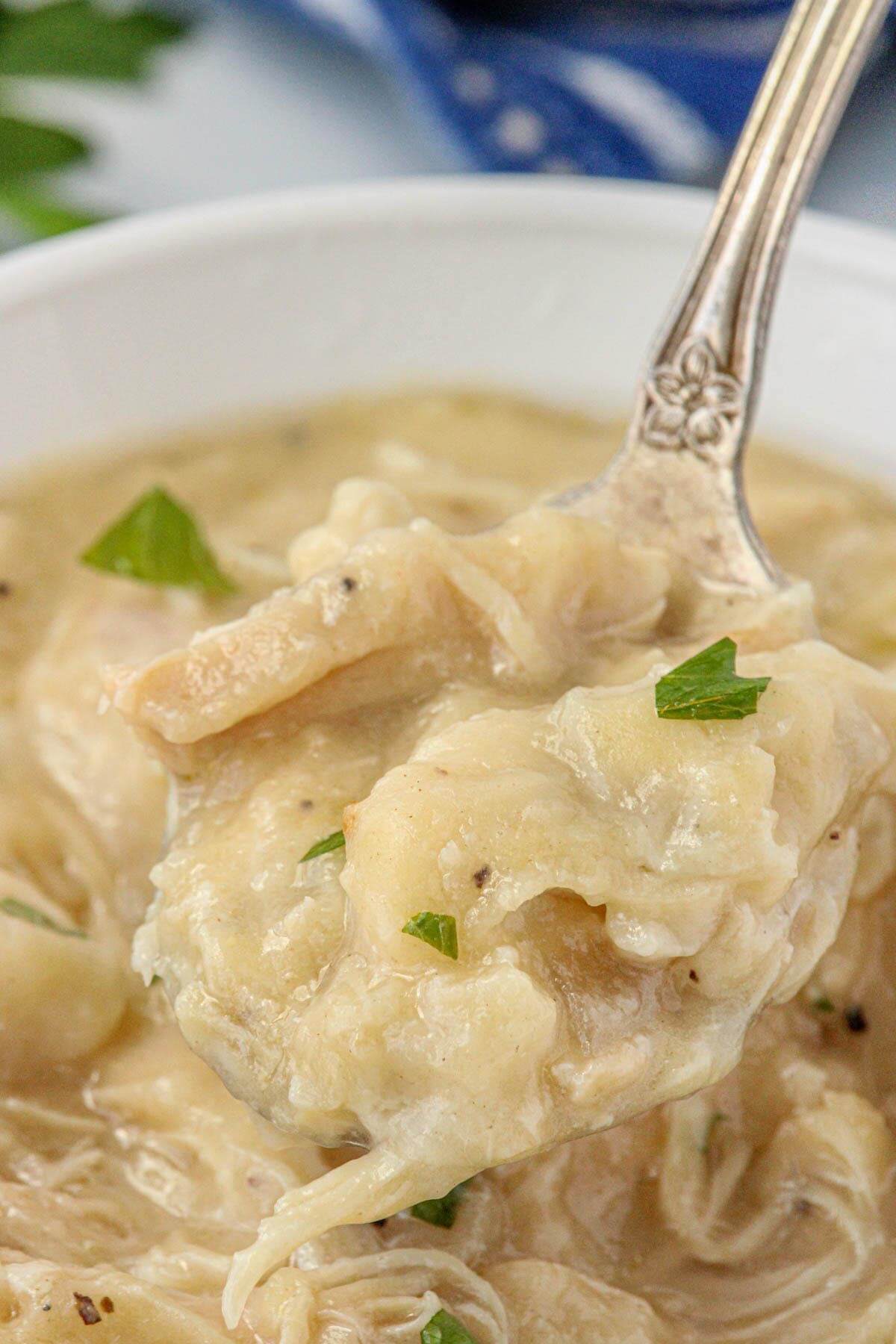 Silver spoon holding a bite of tender chicken and dumplings topped with fresh parsley over a creamy broth.