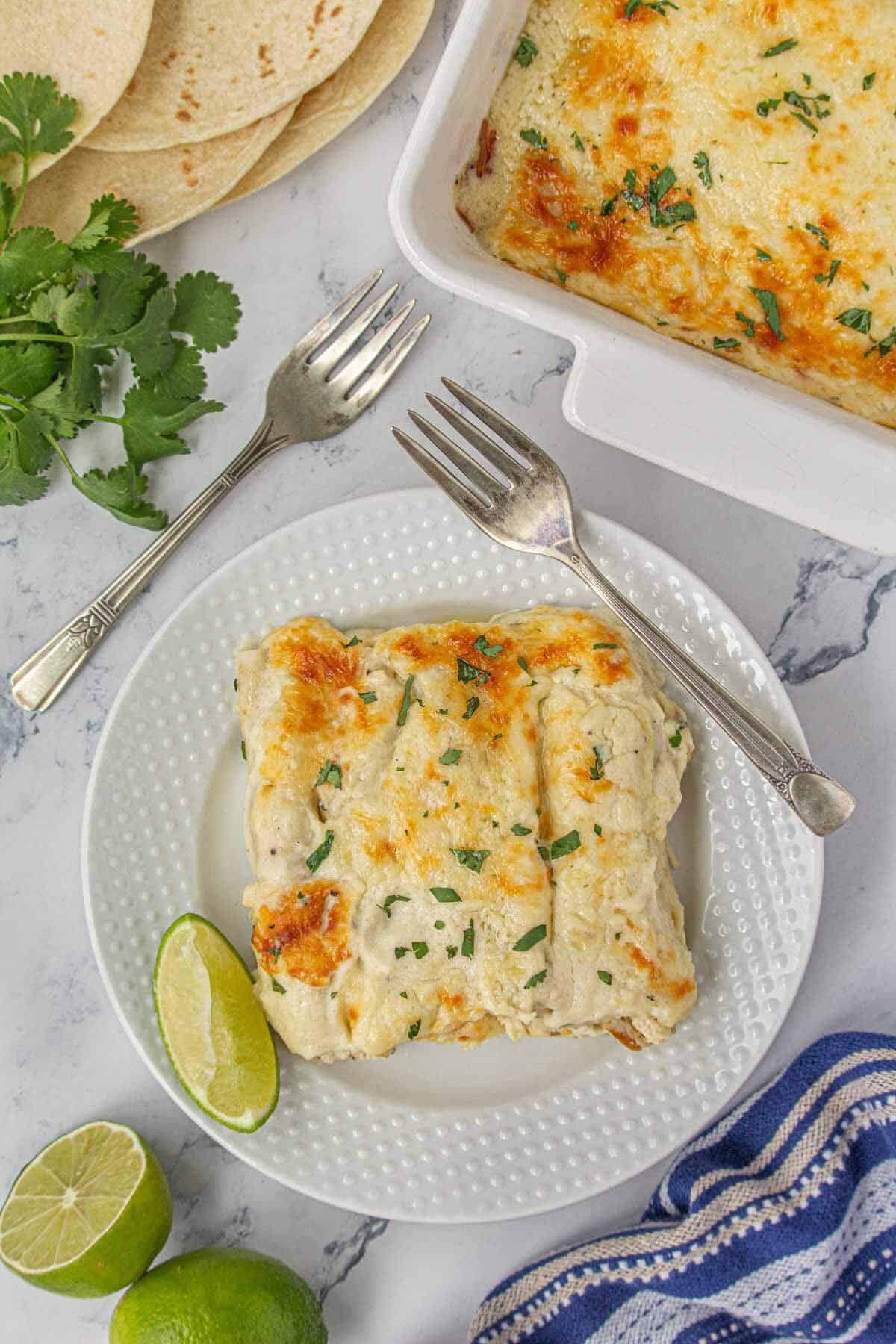 Overhead view of a plated serving of cheesy sour cream chicken enchiladas with lime wedges, fresh cilantro, and a bubbling casserole dish in the background.