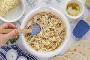 A white mixing bowl filled with shredded chicken, diced green chiles, and shredded cheese being stirred with a blue spatula.