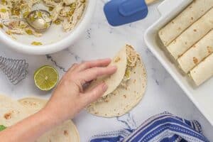 Hand folding a flour tortilla around creamy chicken enchilada filling, with more rolled tortillas lined up in a white baking dish beside it.