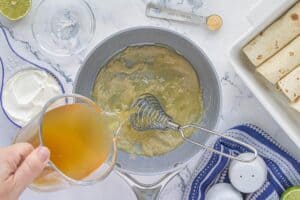 Chicken broth being poured into a roux in a saucepan to make homemade white enchilada sauce, surrounded by sour cream and seasonings.