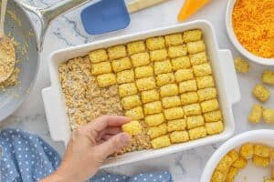 A hand placing frozen tater tots over a beef and cheese mixture in a white casserole dish, assembling the second layer of the casserole.