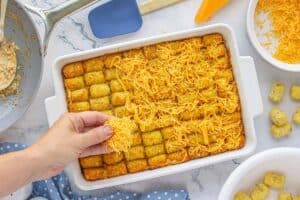 Hand sprinkling shredded cheddar cheese over golden baked tater tots in a white baking dish, final step before returning to the oven.