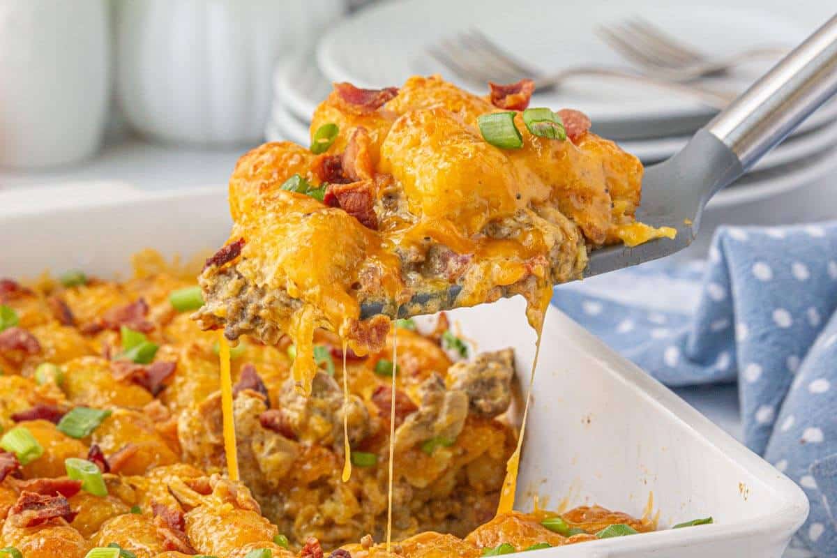 Close-up of a spatula lifting a generous scoop of cheesy bacon cheeseburger tater tot casserole from a white baking dish.