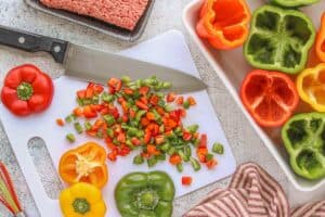 Chopped red, green, and orange bell pepper tops on a cutting board beside a sharp knife and a baking dish filled with hollowed whole peppers ready for stuffing.