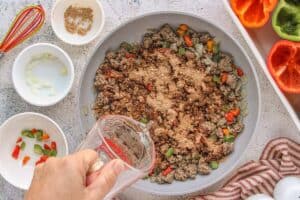 Ground beef sautéing in a skillet with diced peppers and onions while taco seasoning and water are added, surrounded by prep bowls and halved bell peppers.