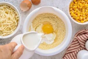 Milk being poured into a mixing bowl filled with Jiffy cornbread mix and an egg, with bowls of shredded cheese and corn in the background ready to be added.