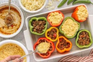 Stuffed bell peppers in a white baking dish, half filled with seasoned beef mixture and others topped with creamy cornbread batter, ready to be baked.